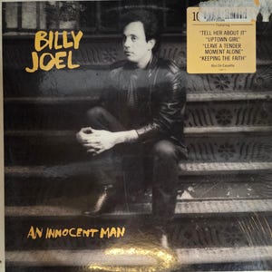 May include: Album cover for Billy Joel's "An Innocent Man." The album art features a seated man in a leather jacket on a staircase. The album title is in gold lettering. A sticker on the upper right lists songs including "Uptown Girl."