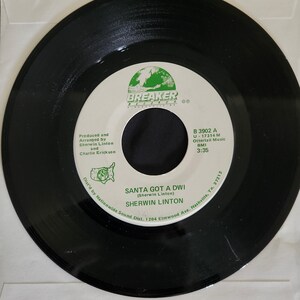 May include: A black and white 45 RPM vinyl record with a white label. The label has a green globe graphic and the text "BREAKER RECORDS". The record title is "SANTA GOT A DWI (Sherwin Linton)" and the artist is "SHERWIN LINTON". The record label also includes the song information: "B 3902 A, U-17314 M, Ottertail Music BMI, 3:35".