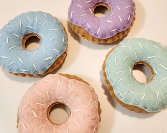May include: Four felt donuts with different colored frosting and white sprinkles. The donuts are light blue, purple, pink, and light green.