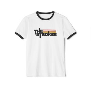 May include: A white ringer t-shirt with black trim on the collar and sleeve cuffs. The chest features the text THE STROKES in black, accompanied by three horizontal stripes in red, orange, and yellow positioned above the word STROKES.