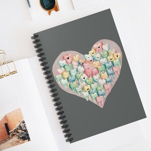 May include: A grey spiral notebook featuring a pink heart design. The heart is filled with colourful cartoon teeth with smiling faces.