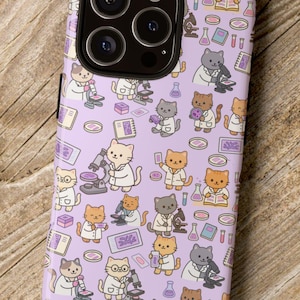 Peut inclure: Une coque de téléphone lavande avec un motif répété de chats de dessin animé en blouses de laboratoire, utilisant des microscopes et lisant des livres. L'étui a une découpe noire pour l'objectif de l'appareil photo et est posé sur une surface en bois.