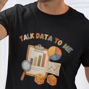 Puede incluir: Camiseta negra con el texto "TALK DATA TO ME" sobre un gráfico de gráficos de estadísticas, gráficos, dados y una lupa. La camiseta la lleva una persona.