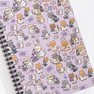 Peut inclure: Un cahier à spirales avec une couverture lavande ornée d'un motif répété de chats de dessin animé en blouses de laboratoire, utilisant des microscopes et des béchers. Le cahier est ouvert sur une surface blanche.