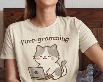 May include: A beige t-shirt with the text "Purr-gramming in Progress" and a cartoon cat using a laptop. A person is wearing the shirt and sitting on a bed with a cat. The cat is white and gray.