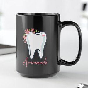May include: Black ceramic mug featuring a white tooth graphic with pink floral accents. The tooth has a pink heart on it. The mug reads "Amanda" in pink lettering.
