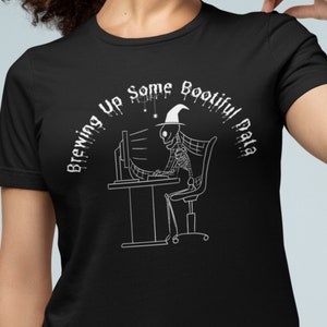 Könnte beinhalten: Schwarzes T-Shirt mit einer weißen Grafik eines Skeletts mit Hexenhut, das vor einem Computer sitzt. Der Text "Brewing Up Some Bootiful Data" ist über dem Skelett angeordnet.