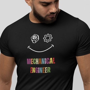 Może przedstawiać: Czarny T-shirt z białą uśmiechniętą twarzą wykonaną z nakrętki i zębatki. Tekst "MECHANICAL ENGINEER" jest napisany pod twarzą w kolorach tęczy.