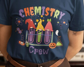Può includere: T-shirt blu navy con la scritta "CHEMISTRY CREW" a lettere colorate. La maglietta presenta una grafica di tre gnomi in provette, insieme a illustrazioni a tema scientifico e una zucca di Halloween. È visibile una cintura marrone.