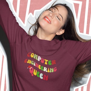 May include: A maroon sweatshirt with the text "COMPUTER ENGINEERING QUEEN" in colorful, bubble letters. The sweatshirt is long-sleeved and has a crew neck. The background is a pink and white geometric design.