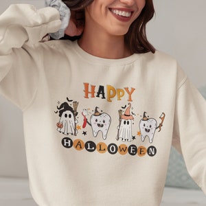 Puede incluir: Una sudadera de color crema con un gráfico de cuatro personajes de dibujos animados vestidos de brujas. Los personajes son un fantasma, dos dientes y otro fantasma. El texto "HAPPY HALLOWEEN" está impreso debajo de los personajes.