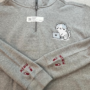 May include: Gray quarter-zip sweatshirt with a cat graphic on the front. The cat is sitting in front of a laptop. The sleeves have the word "MEOW" and paw prints embroidered in maroon.