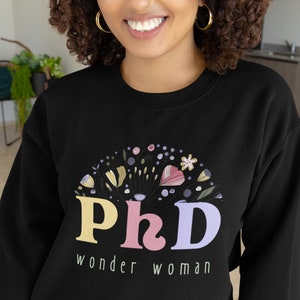 May include: Black sweatshirt with a floral design and the text "PhD wonder woman" in pastel colors. The design includes flowers and leaves. The model is smiling and wearing gold hoop earrings.