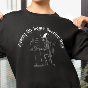 Könnte beinhalten: Schwarzes Sweatshirt mit dem Text "Brewing Up Some Bootiful Data" und einer Skelettgrafik. Das Skelett trägt einen Hexenhut und sitzt an einem Computer. Die Person auf dem Bild trägt blaue Shorts.