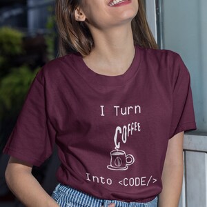 Może przedstawiać: Bordowy t-shirt z białym napisem "I turn coffee into <CODE/>". Tekst jest uzupełniony rysunkiem filiżanki kawy z unoszącą się parą.