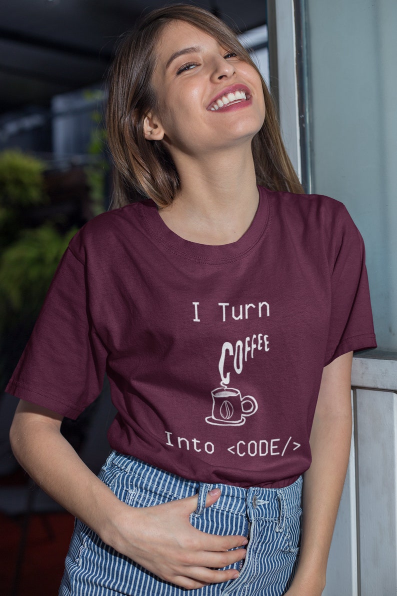 Może przedstawiać: Bordowy t-shirt z białym napisem "I turn coffee into <CODE/>". Tekst jest uzupełniony rysunkiem filiżanki kawy z unoszącą się parą.
