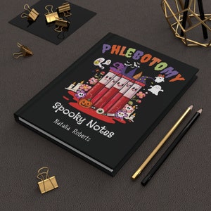 May include: Black journal with the words "Phlebotomy Spooky Notes" and the name "Natalia Roberts." The cover features a Halloween-themed illustration with blood vials, ghosts, and a pumpkin. Gold and black pencils and paper clips are also visible.
