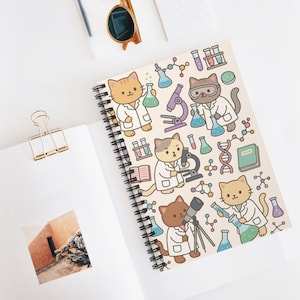 May include: Spiral-bound notebook with a repeating pattern of cartoon cats in lab coats, beakers, microscopes, and scientific diagrams. The notebook is open on a white surface with a gold paper clip and sunglasses.