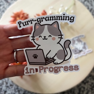 Op de afbeelding: Een sticker met een cartoonkat die op een laptop werkt. De kat is wit en grijs, met de tekst "Purr-gramming in Progress". De sticker wordt door een hand vastgehouden.