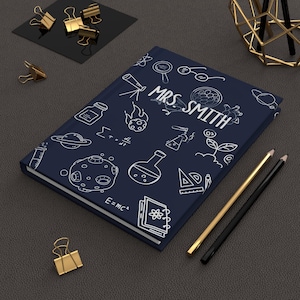 May include: A navy blue journal with white science-themed doodles and the name "MRS SMITH" in white. The journal is next to gold and black pencils and gold binder clips on a dark gray surface.