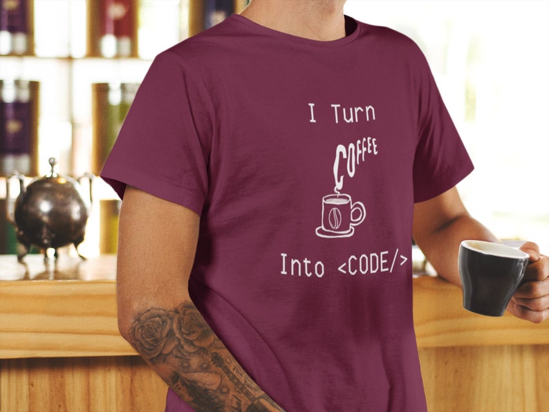 Może przedstawiać: Bordowy t-shirt z nadrukiem "I Turn Coffee Into <CODE/>" na przodzie. Tekst jest biały i przedstawia stylizowaną filiżankę kawy z unoszącą się parą.