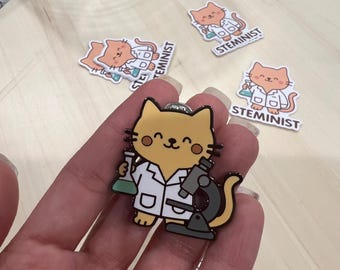 Op de afbeelding: Een gele kattenemaille pin met een labjas, een beker en een microscoop. Op de pin staat het woord "STEMINIST" gedrukt. Extra pins zijn op de achtergrond verspreid.