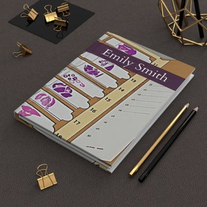 Könnte beinhalten: Ein personalisiertes Notizbuch mit dem Namen "Emily Smith" auf einem lila Band. Das Notizbuch hat ein einzigartiges Design mit Illustrationen und einem nummerierten Raster. Goldene und schwarze Bleistifte und goldene Büroklammern sind ebenfalls vorhanden.