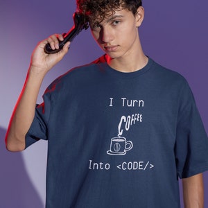 Może przedstawiać: Koszulka w kolorze granatowym z białym nadrukiem, na kt&oacute;rym widnieje napis "I Turn Coffee Into <CODE/>" wraz z kresk&oacute;wkową filiżanką kawy i parą.