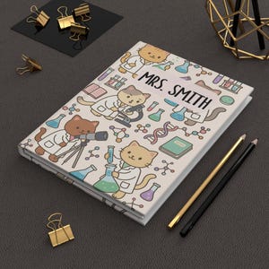 May include: A cream-coloured notebook with a cover illustrated with cartoon cats in lab coats and science symbols. The notebook is labelled "MRS. SMITH". Gold and black pencils and paper clips are also visible.