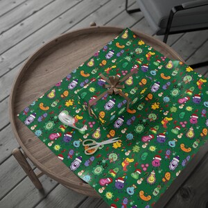 May include: A wrapped gift box with a brown ribbon sits on green wrapping paper. The paper and box feature a repeating pattern of colourful cartoon microbes wearing Santa hats and scarves. A pair of scissors and a roll of tape are also on the paper.