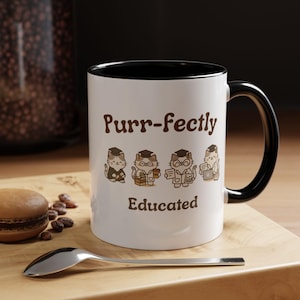 Peut inclure: Mug en céramique blanche avec intérieur et anse noirs. Le mug présente le texte "Purr-fectly Educated" avec des illustrations de chats de dessins animés portant des chapeaux de graduation et des lunettes. Une cuillère et un macaron sont sur une surface en bois.