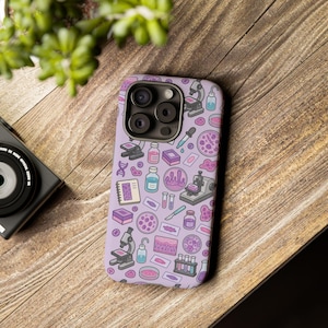 May include: A light purple smartphone case featuring a repeating pattern of laboratory equipment including microscopes, test tubes, petri dishes, DNA strands, scalpels, and various vials, all rendered in a colorful, illustrated graphic style.