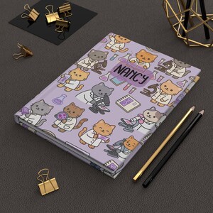 Peut inclure: Un journal à couverture rigide avec une couverture lavande ornée de chats de dessins animés en blouses de laboratoire, microscopes et béchers. Le nom "NANCY" est imprimé sur la couverture. Des crayons et des trombones dorés et noirs sont également visibles.