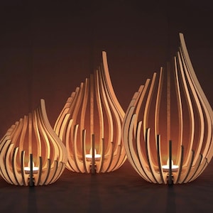 Puede incluir: Tres candelabros de madera con forma de lágrima. Los soportes están hechos de listones de madera verticales, con una vela encendida dentro de cada uno. La madera es de color marrón claro y el fondo es marrón oscuro.