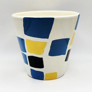May include: A white ceramic vase with a geometric pattern of blue, yellow, and black squares. The vase has a cylindrical shape and a slightly flared top. The design is evenly spaced around the vase.