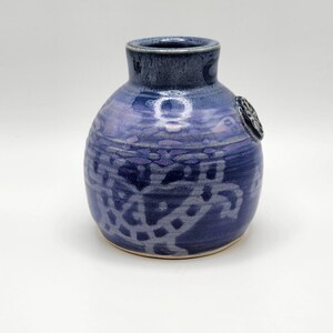 May include: A small, handcrafted ceramic vase in shades of blue. The vase has a rounded body with a narrow neck and a dark blue rim. The surface features a textured, patterned design and a small, circular emblem.