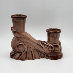 May include: A brown ceramic candle holder with two cylindrical openings. The holder has a decorative, ornate design with a curved base and a textured surface. The candle holder is designed to hold two candles.