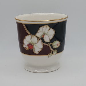 May include: A white ceramic cup with a floral design. The cup features a white orchid design with red centers and green leaves. The top portion is black and burgundy, with a gold trim. The base is white.