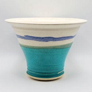 May include: A handcrafted ceramic vase with a flared shape. The vase features a white top, a band of blue and green stripes, and a turquoise lower section. The vase is approximately 8 inches tall and 8 inches wide.