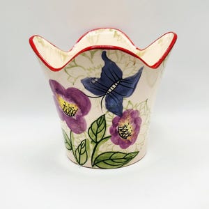 May include: A ceramic vase with a tulip-shaped design. The vase is painted with purple flowers, green leaves, and a blue butterfly. The rim is red, and the background is off-white.