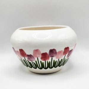May include: A white ceramic bowl with a rounded shape. The bowl is decorated with a hand-painted floral design featuring pink, red, and purple tulips with green stems and leaves. The bowl is likely used for decorative purposes.