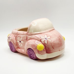 May include: A pink ceramic car-shaped planter with white daisy accents. The planter has purple wheels and yellow headlights. The top of the car is off-white, and the interior is hollow for planting flowers or small plants.
