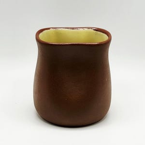 May include: A small, brown ceramic vase with a rounded body and a slightly flared rim. The interior is a light yellow color, contrasting with the exterior. The vase is likely handmade and suitable for small floral arrangements or decorative use.