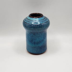 May include: A small, handcrafted ceramic vase with a unique shape. The vase features a vibrant teal glaze with darker blue accents and a brown rim. The design includes horizontal ridges around the top, adding texture to the piece. The vase is approximately 5 inches tall.