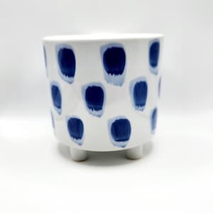 May include: White ceramic planter with a pattern of navy blue brushstrokes. The small, round planter sits on three short legs. A decorative container for plants, flowers, or other items.