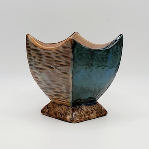 May include: A ceramic vase with a square base and a flared, curved top. The vase is divided into two sections: one with a textured, brown finish and the other with a smooth, blue-green glaze. The interior is a light brown color.