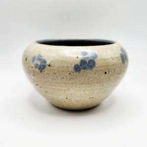 May include: A small, round ceramic bowl with a speckled, beige exterior and a dark interior. The bowl is decorated with blue floral patterns. The bowl is sitting on a white surface.