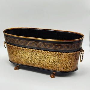 May include: An oval-shaped metal basin with a black interior and a gold-colored exterior featuring an intricate black filigree pattern. The basin has a gold rope-style trim, two side handles, and four small claw-shaped feet at the base.