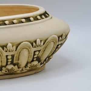 May include: Cream-colored ceramic planter with an oval shape. The planter features a decorative design with raised floral and scrollwork details in a darker, muted green color. The rim is accented with a series of small, raised dots.