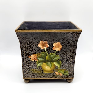 May include: A square, dark brown planter with a distressed, crackled finish. The planter features a hand-painted floral design with orange flowers and green leaves in a yellow pot. The planter sits on four small, round feet.